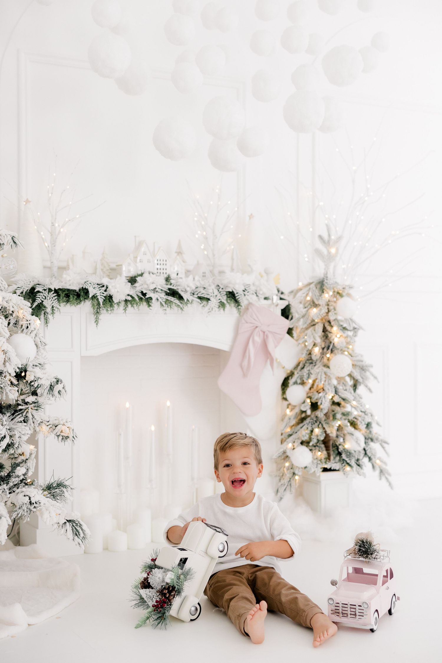 Child playing with pink toy car in white studio with flocked Christmas trees and holiday decor.