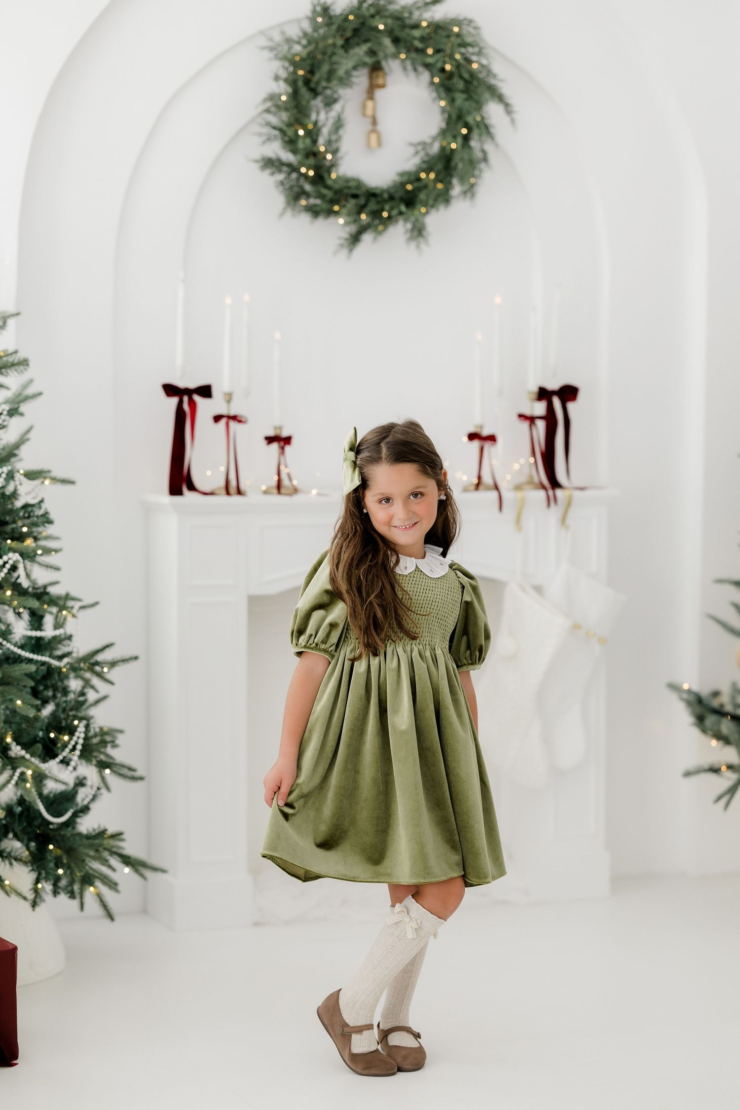 Young girl in green dress poses by Christmas tree and decorated fireplace with wreath and candles.