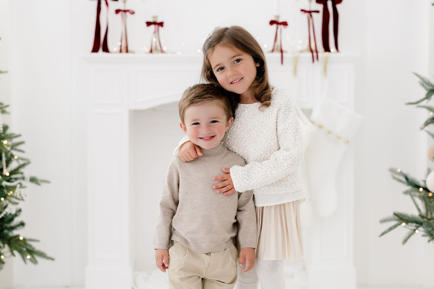 Two young children in white and beige outfits pose together in front of a decorated fireplace during Christmas.