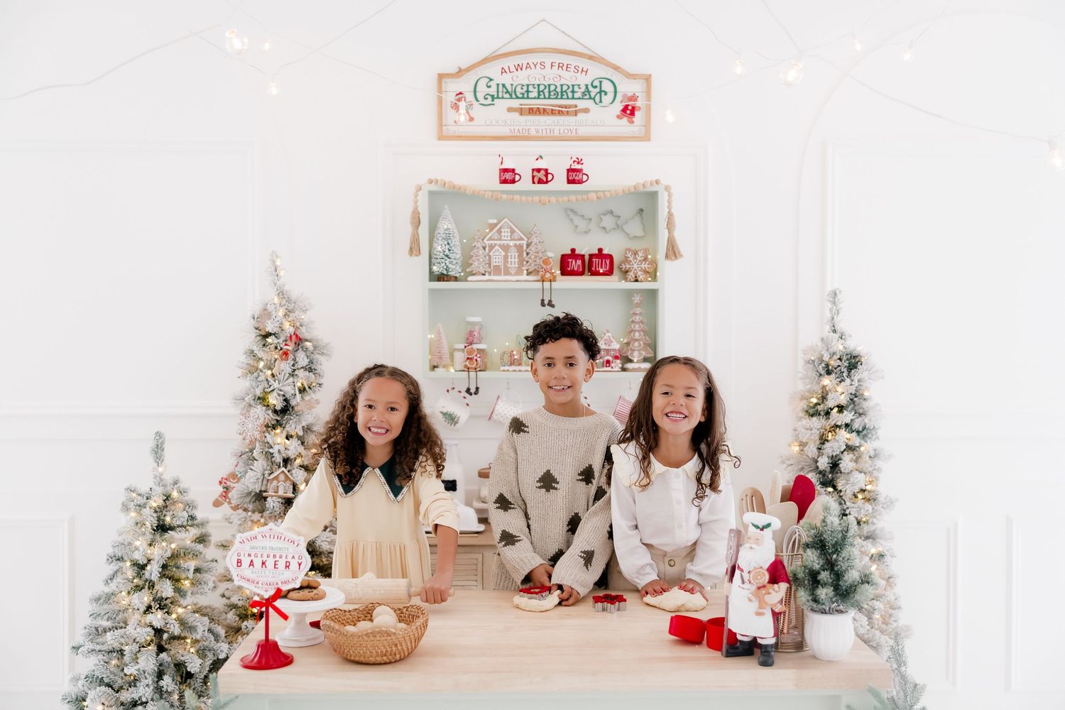 Three children smile at a festive gingerbread-themed baking station surrounded by Christmas trees and holiday decor.