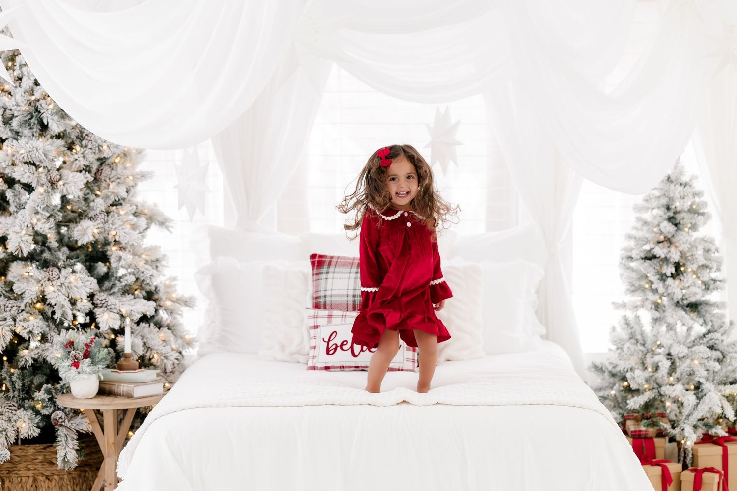 Young girl in red dress jumping on white bed surrounded by snow-flocked Christmas trees with gifts.