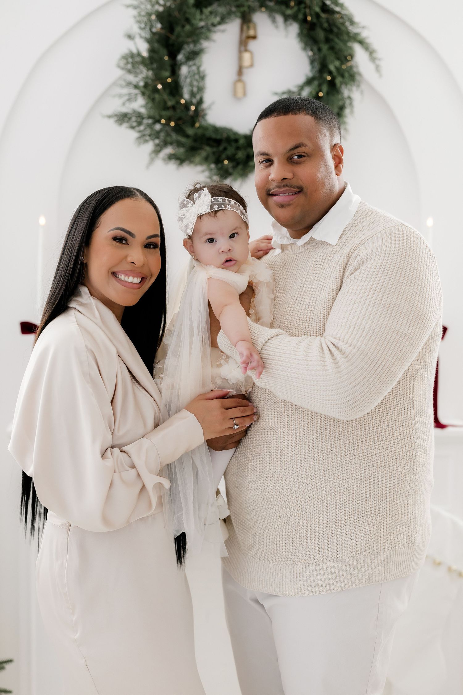 Family portrait with parents holding baby girl in white outfits before Christmas wreath backdrop.