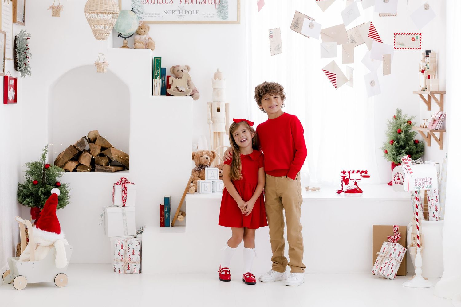 Two children in red holiday outfits pose together in a festive white Christmas-themed photo studio with gifts and decorations.