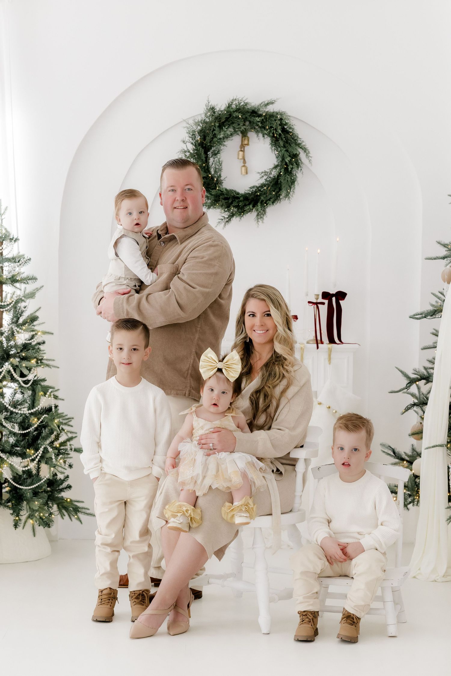 Family of six poses for a festive Christmas photo in a white studio with holiday decor, wreaths, and Christmas trees.