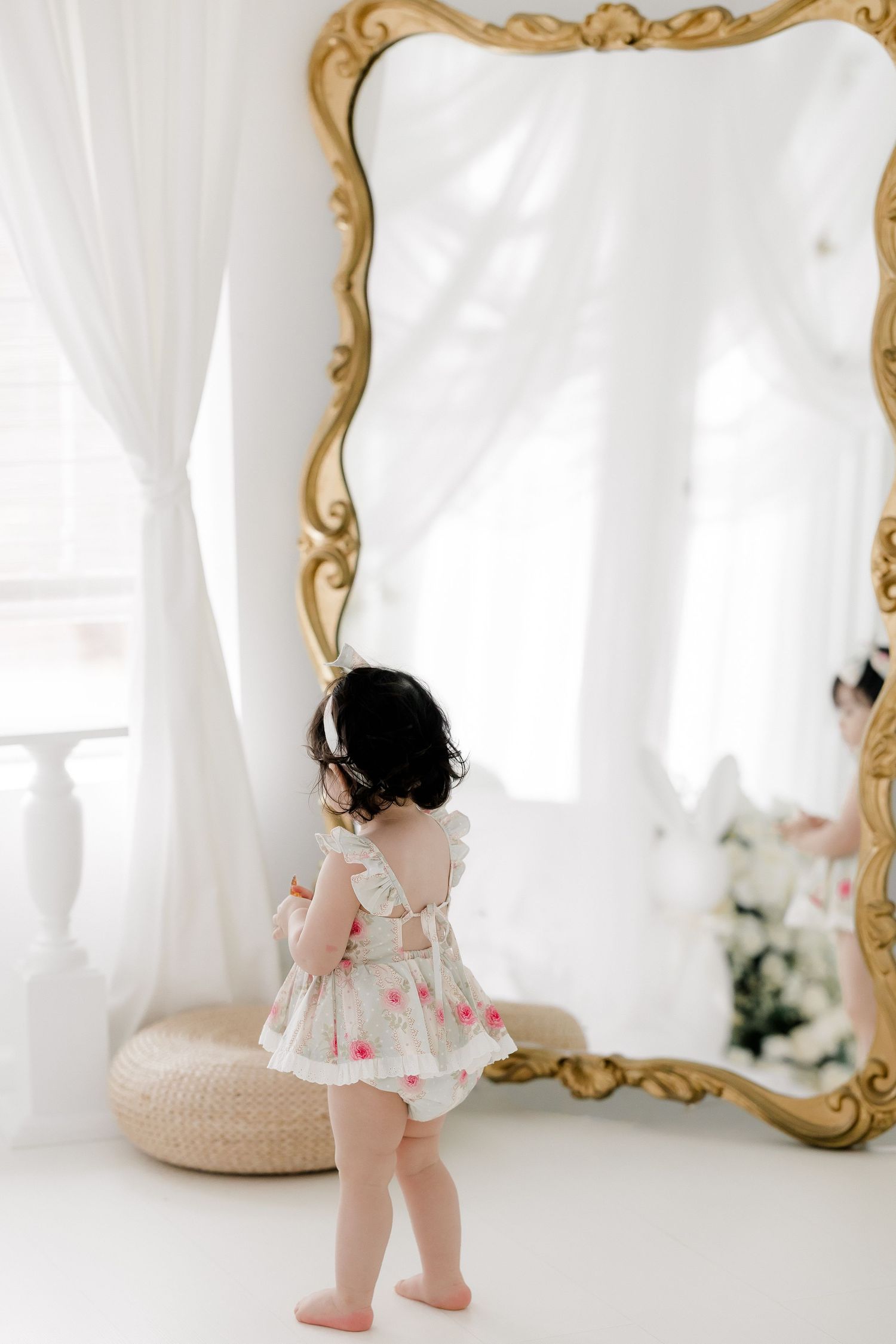 Baby in white dress and bloomers crawls and plays in elegant white studio setting with ornate gold mirror.