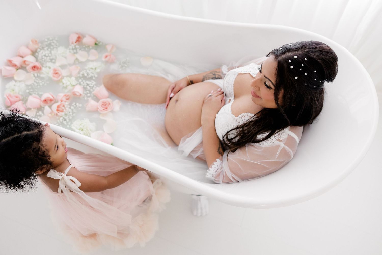 Romantic maternity pose with floral elements and soft pink accents on white background.