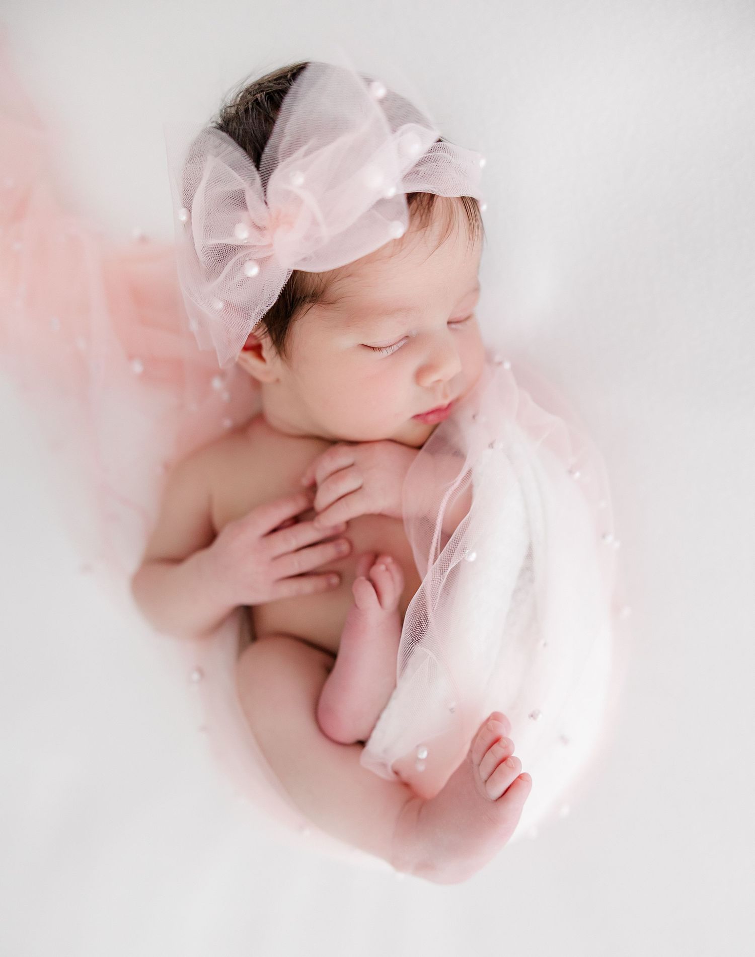 Newborn baby with pink bow headband wrapped in soft tulle on white background.
