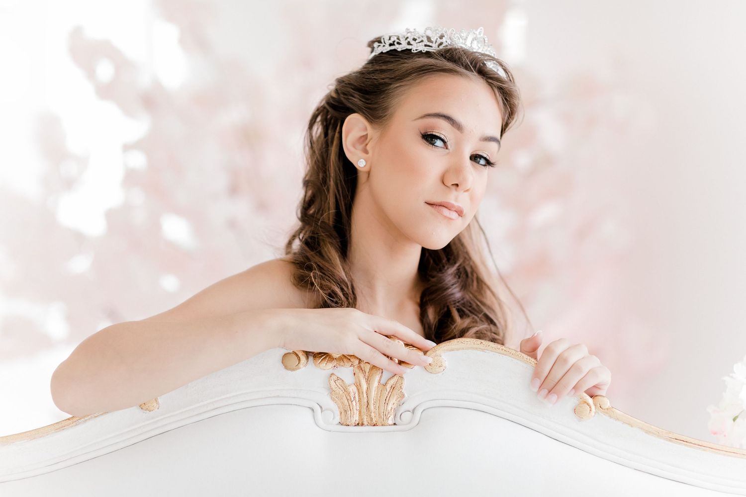 A bride in a white dress and tiara leans gracefully over an elegant white chair with soft pink bokeh in the background.