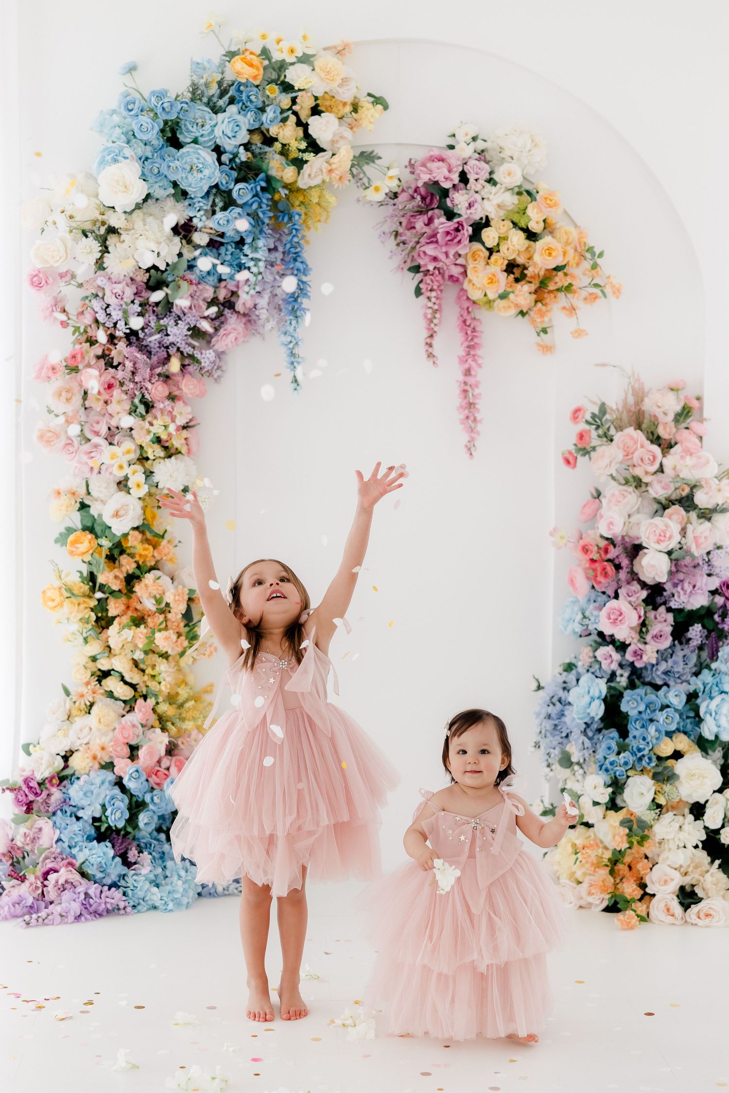 Two small children in pink tulle dresses play under a floral arch with pastel blue, pink, and yellow blooms against white wall.