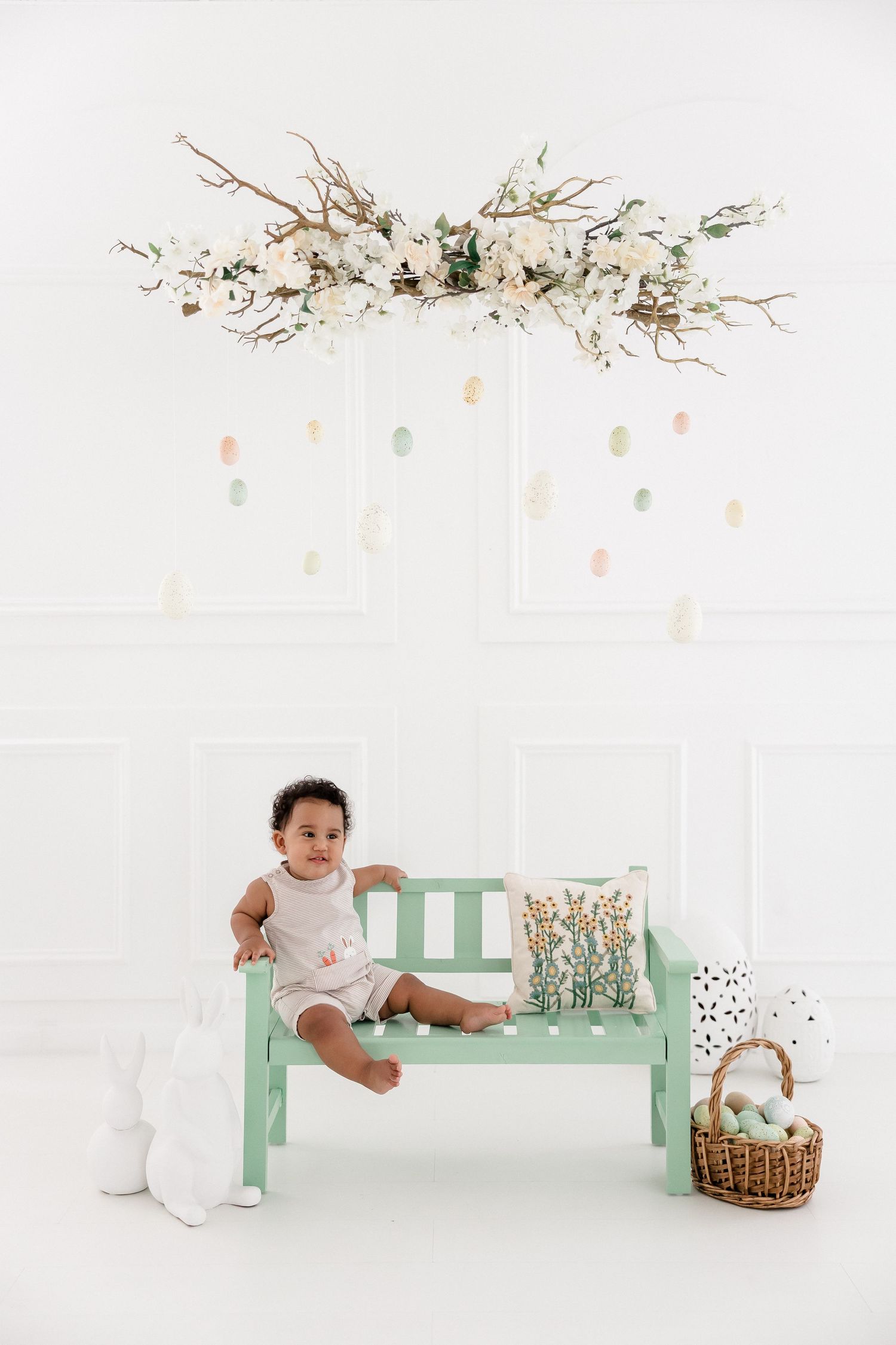 A baby in white clothing sits and plays on a mint green bench with a wicker basket and floral decor in a bright studio setting.