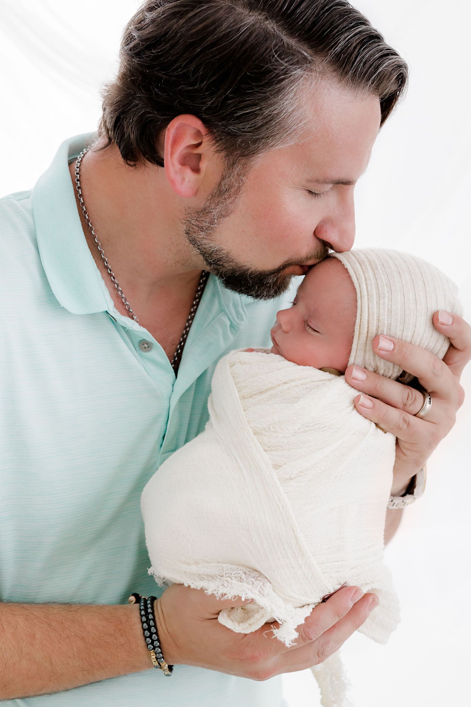 A bearded man in a mint green polo shirt tenderly kisses a newborn baby wrapped in a cream-colored blanket.