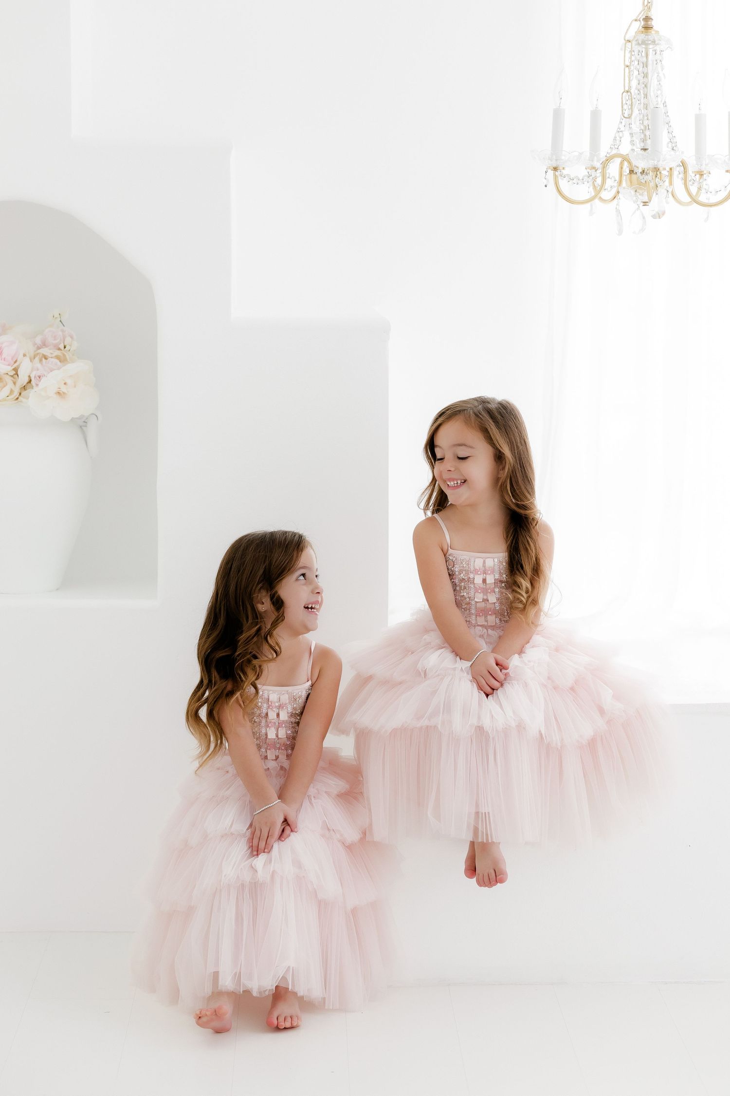 Two young girls in matching pink tulle dresses sit and laugh together in an elegant white room with a chandelier.