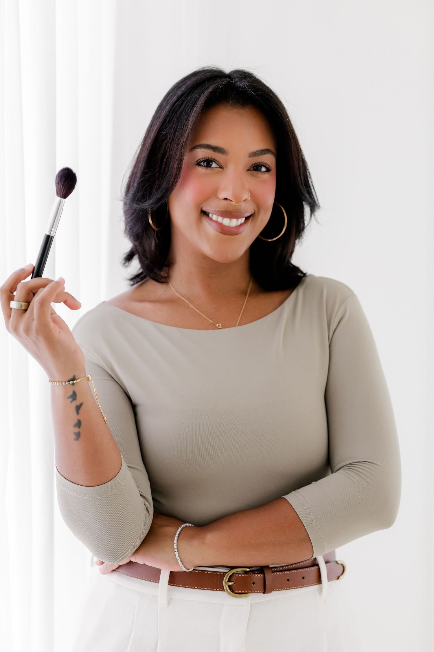 Professional beauty portrait of someone holding a makeup brush against white background, wearing beige top and brown belt.
