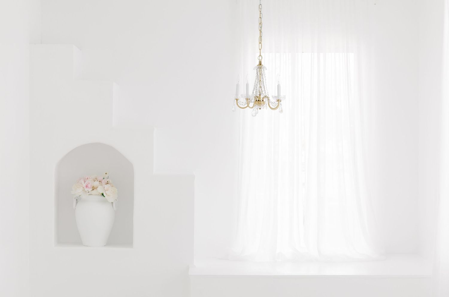 A bright white minimalist room with elegant chandelier and window creating a serene atmosphere.