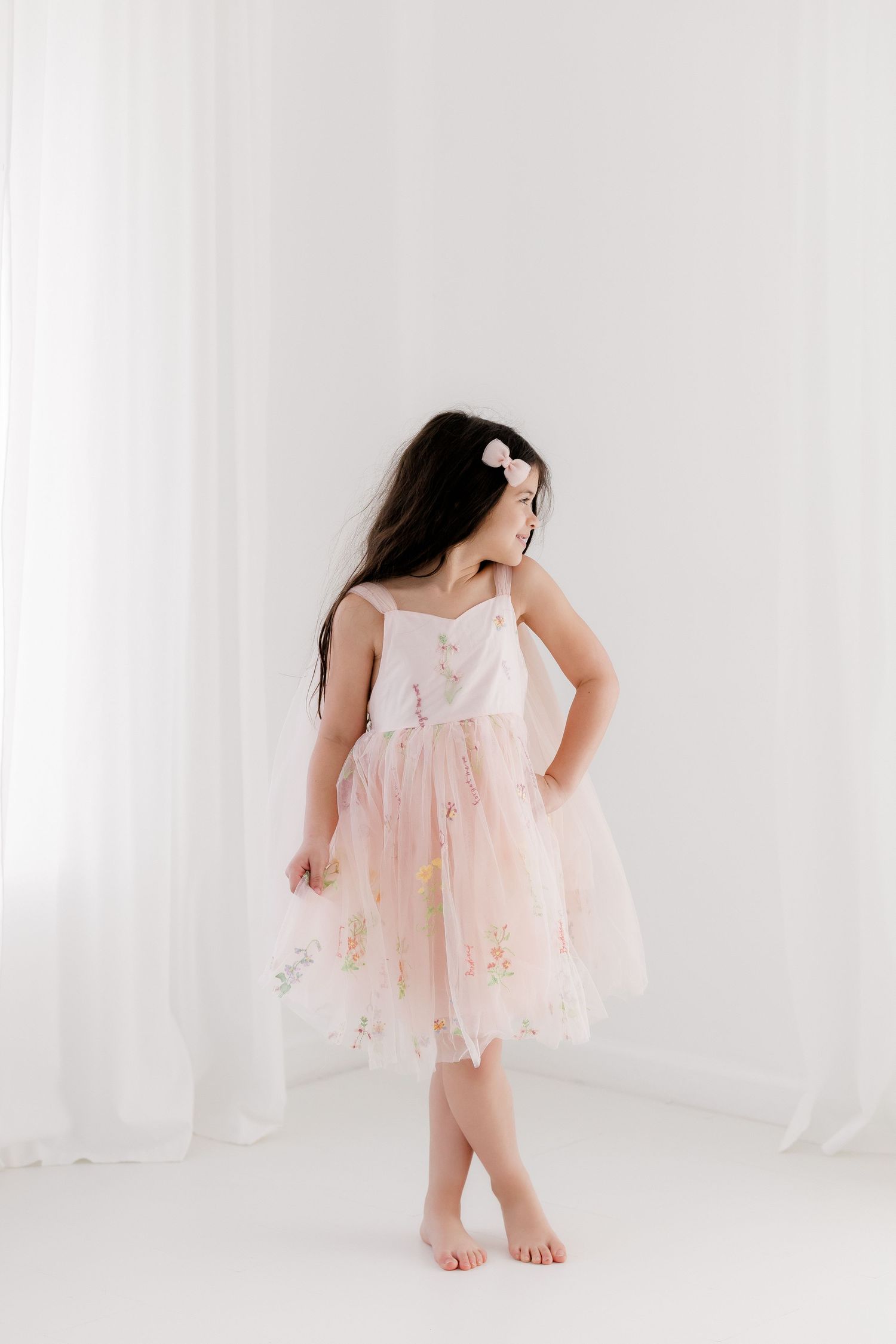Young girl in pink floral tulle dress with hair bow poses barefoot in bright white studio.