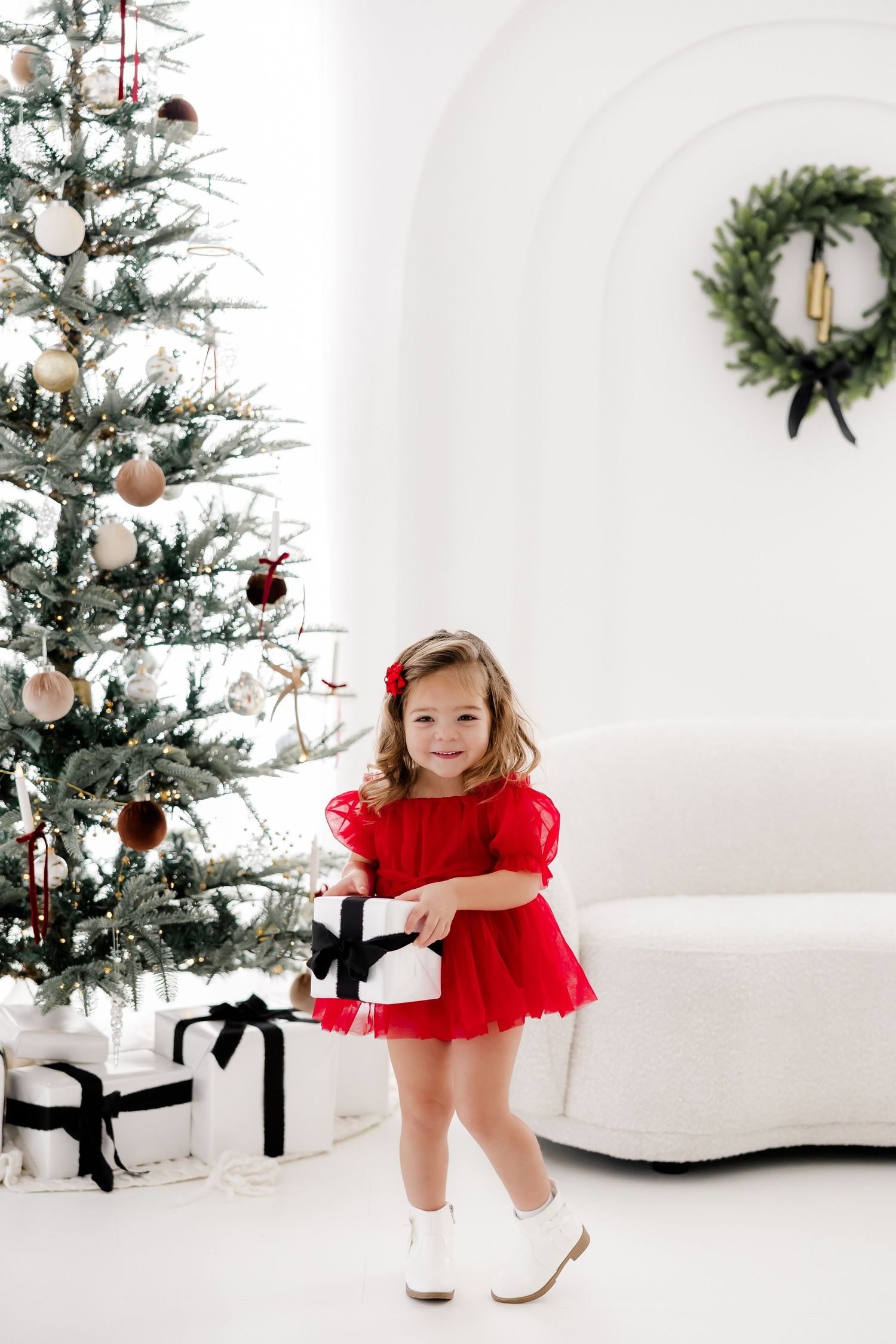 Child in red dress twirls and dances near decorated Christmas tree in white studio setting.