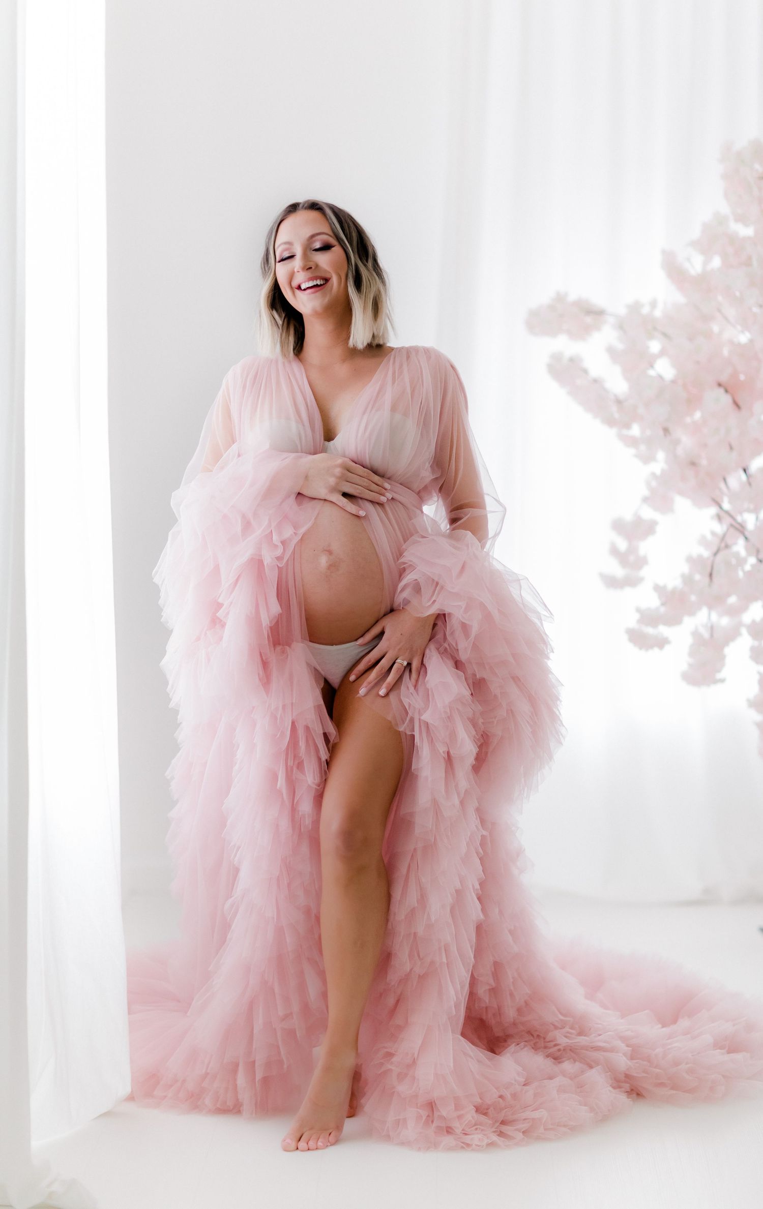 Dreamy maternity photos with figure in flowing pink tulle gown against white backdrop.