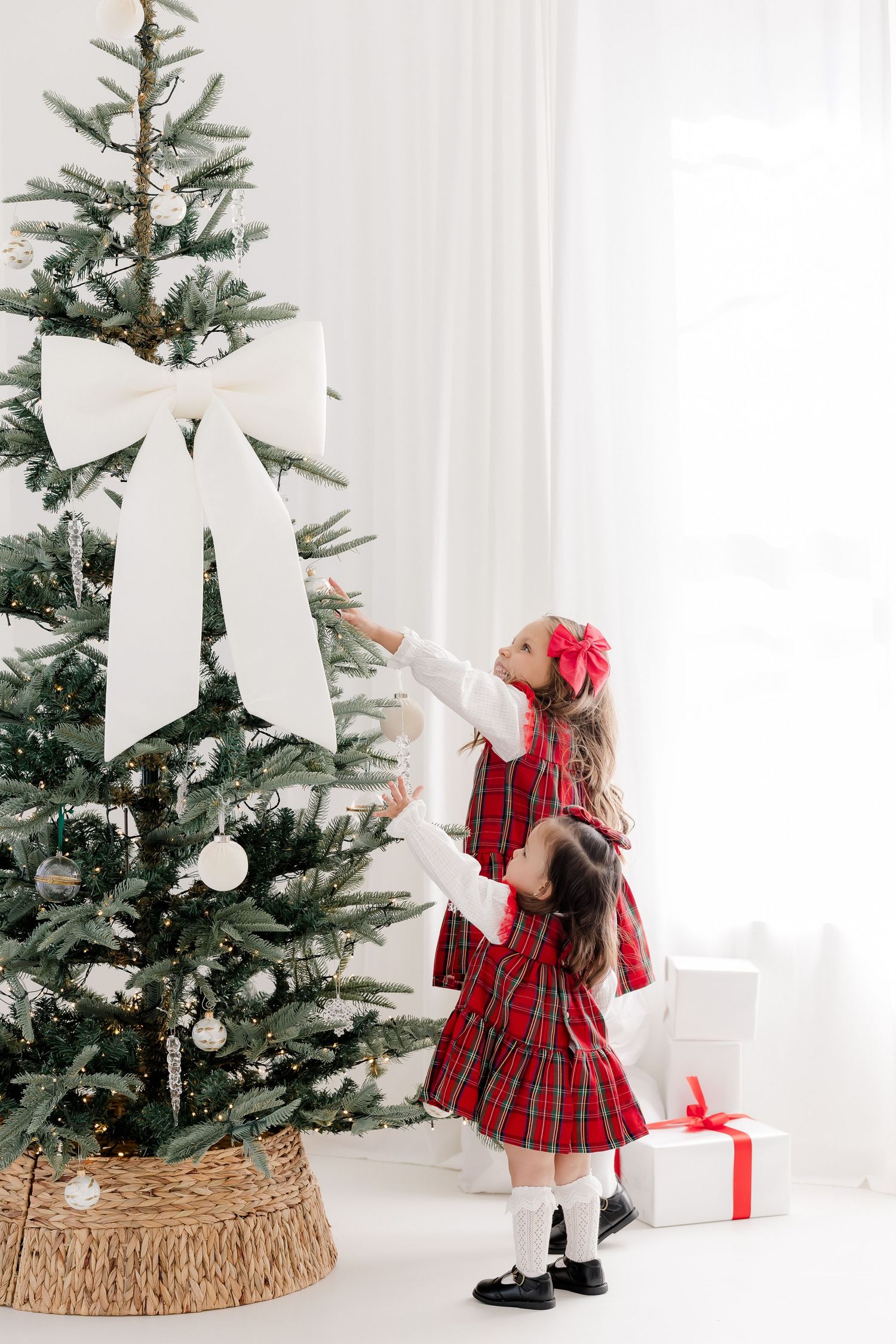 Two girls in red plaid dresses decorating a Christmas tree with white ornaments and bow in a bright white room.