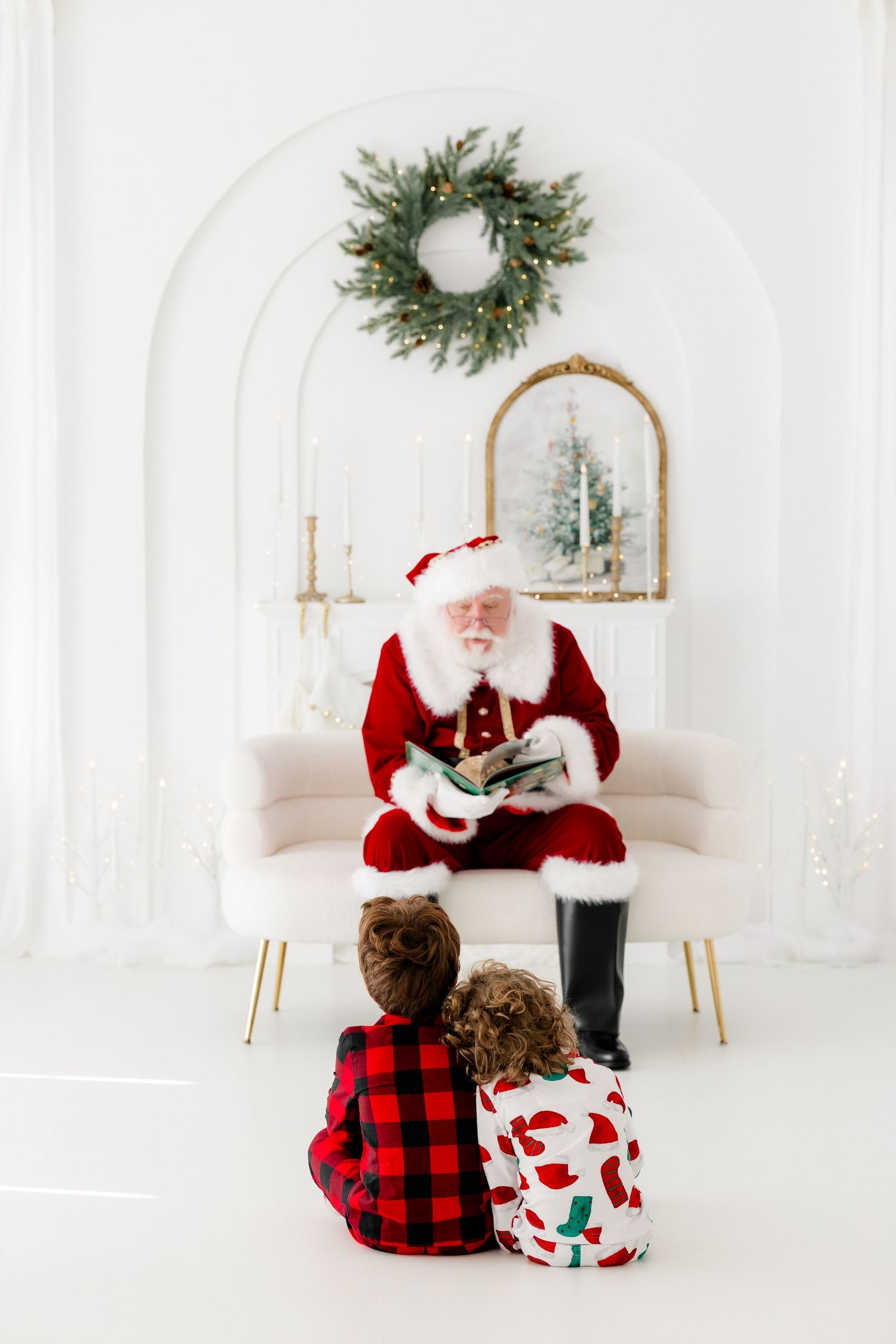 Santa Claus reads a Christmas story to two children in festive pajamas in a white decorated holiday room.