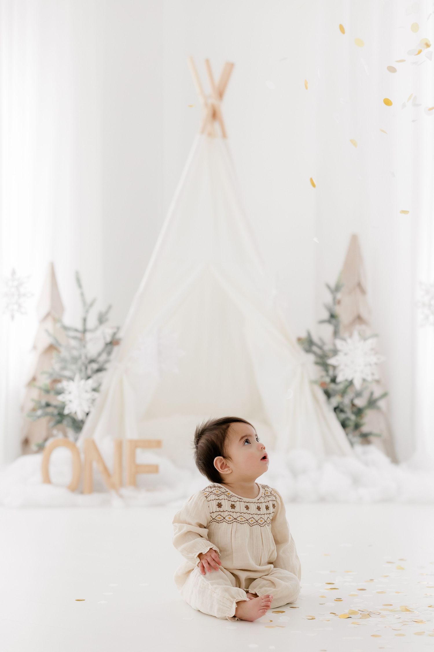 A white teepee tent setup with 'ONE' letters and floral decorations creates a dreamy first birthday photoshoot setting.