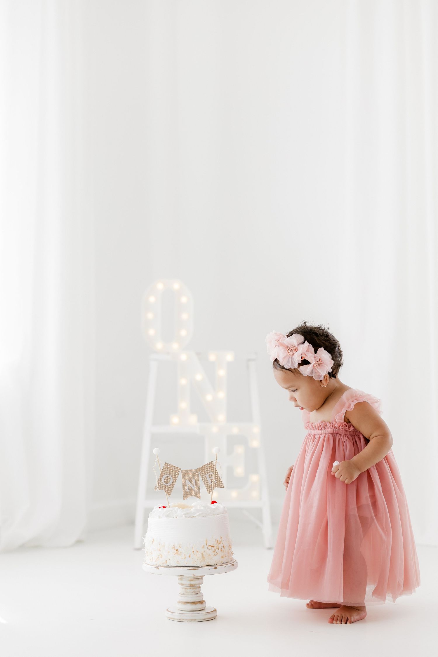 Pink tulle dress cake smash photography session with white backdrop and delicate floral decorations for first birthday celebration.