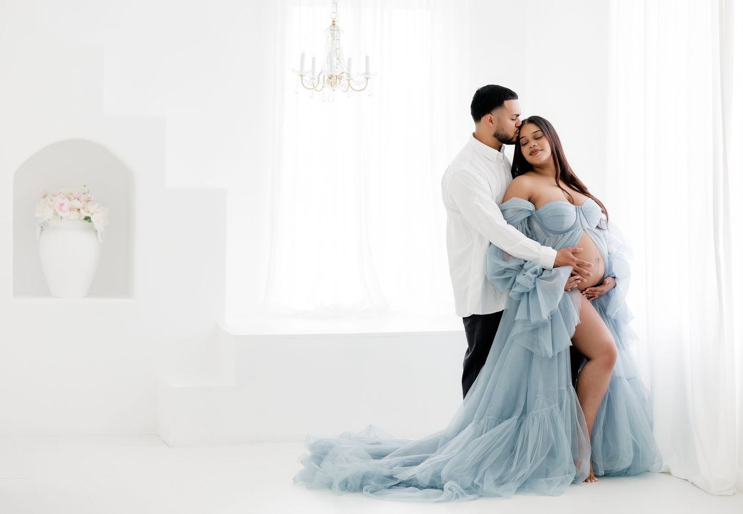 A romantic maternity photoshoot in a light blue tulle gown against a white backdrop.