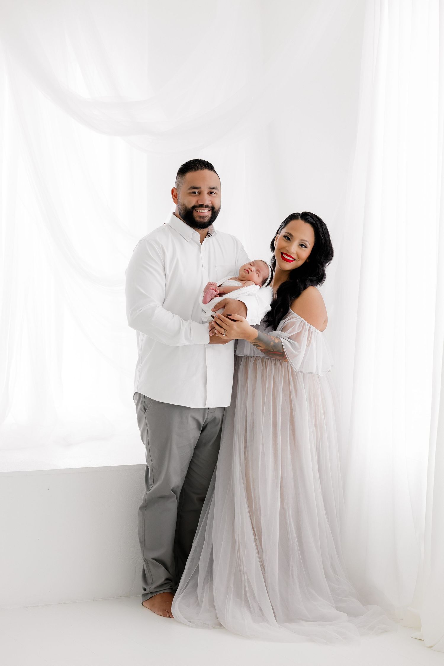 A couple in white and grey attire poses with their newborn baby against a bright white studio backdrop.
