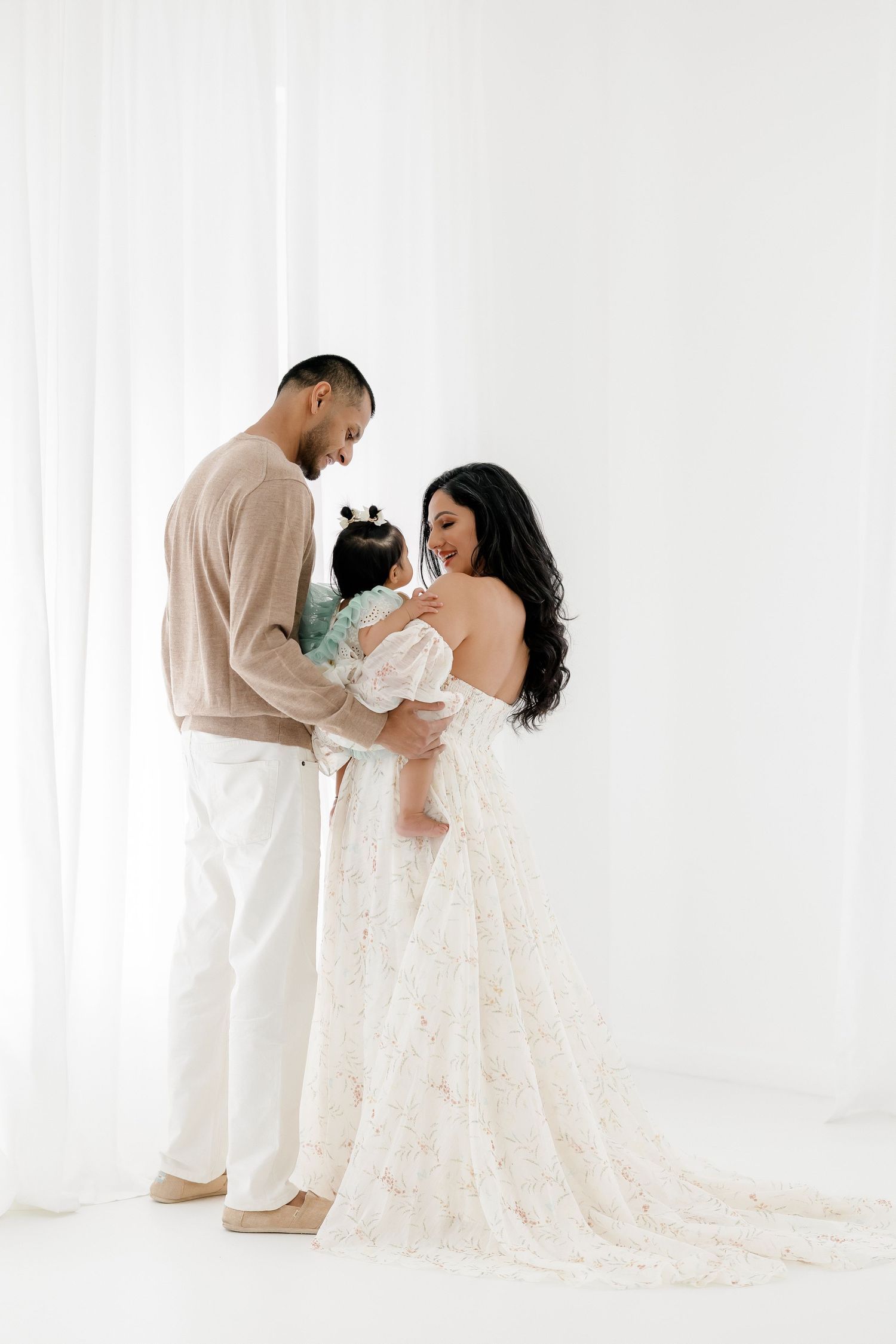 A couple in white outfits shares tender moments with their newborn baby in a bright studio setting.