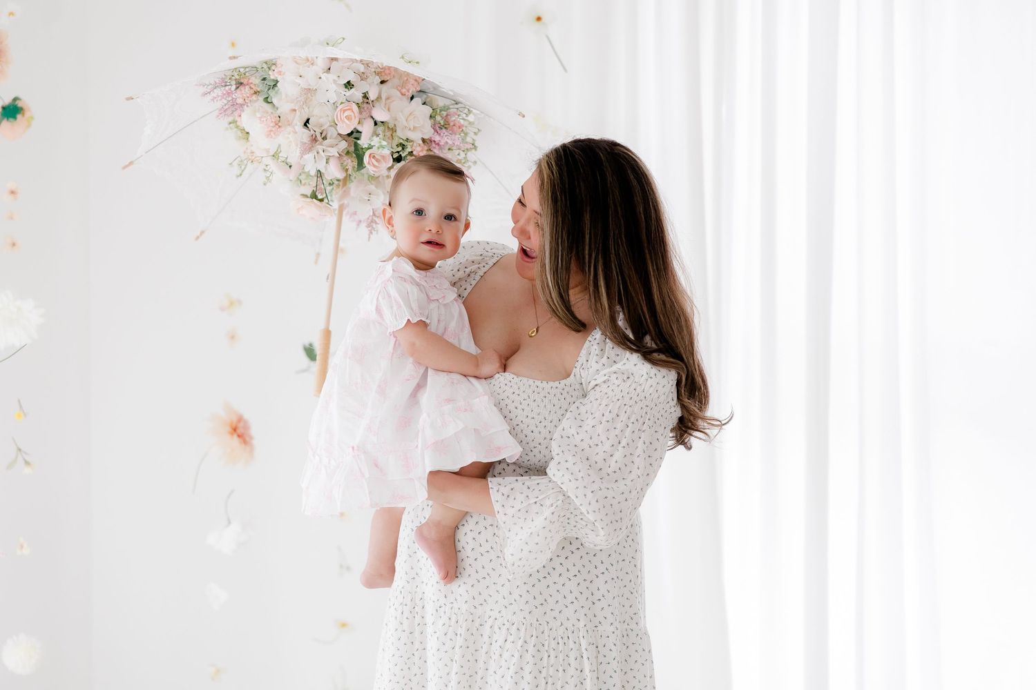 A parent wearing a white dress lifts and plays with a joyful baby in a bright, airy room.