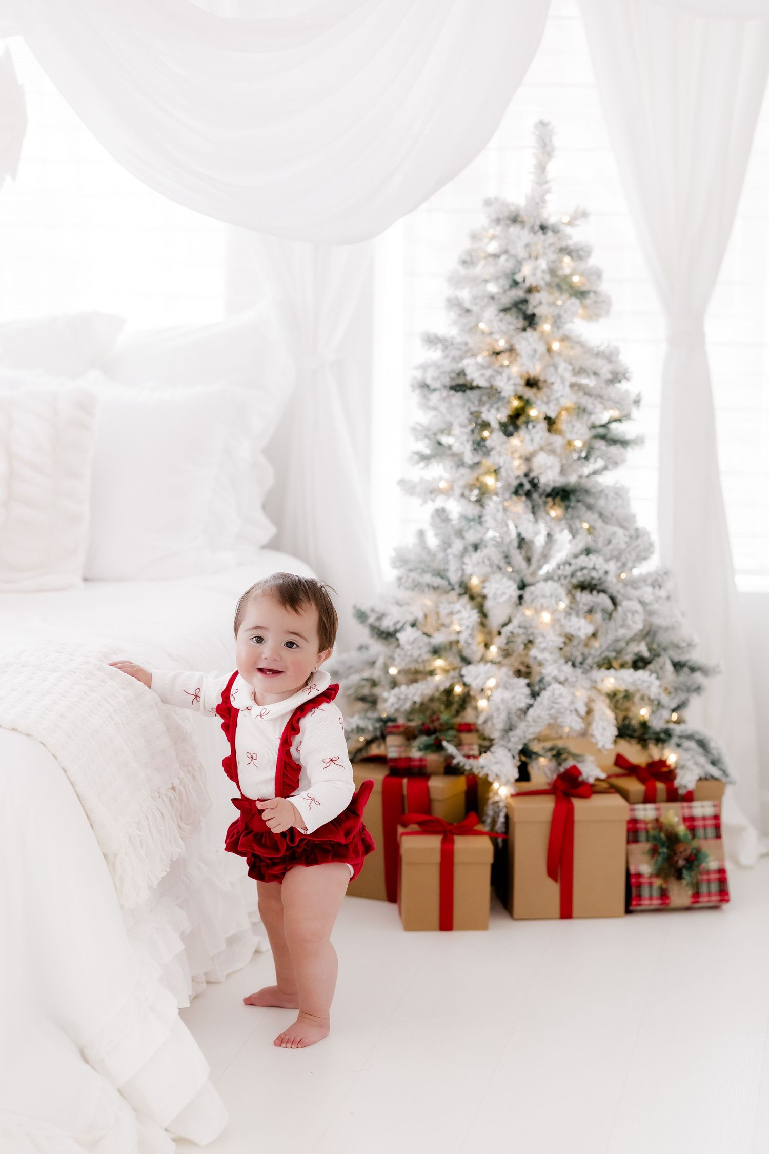 Baby in red Christmas outfit stands by a white flocked tree with gift boxes in a bright white holiday bedroom.