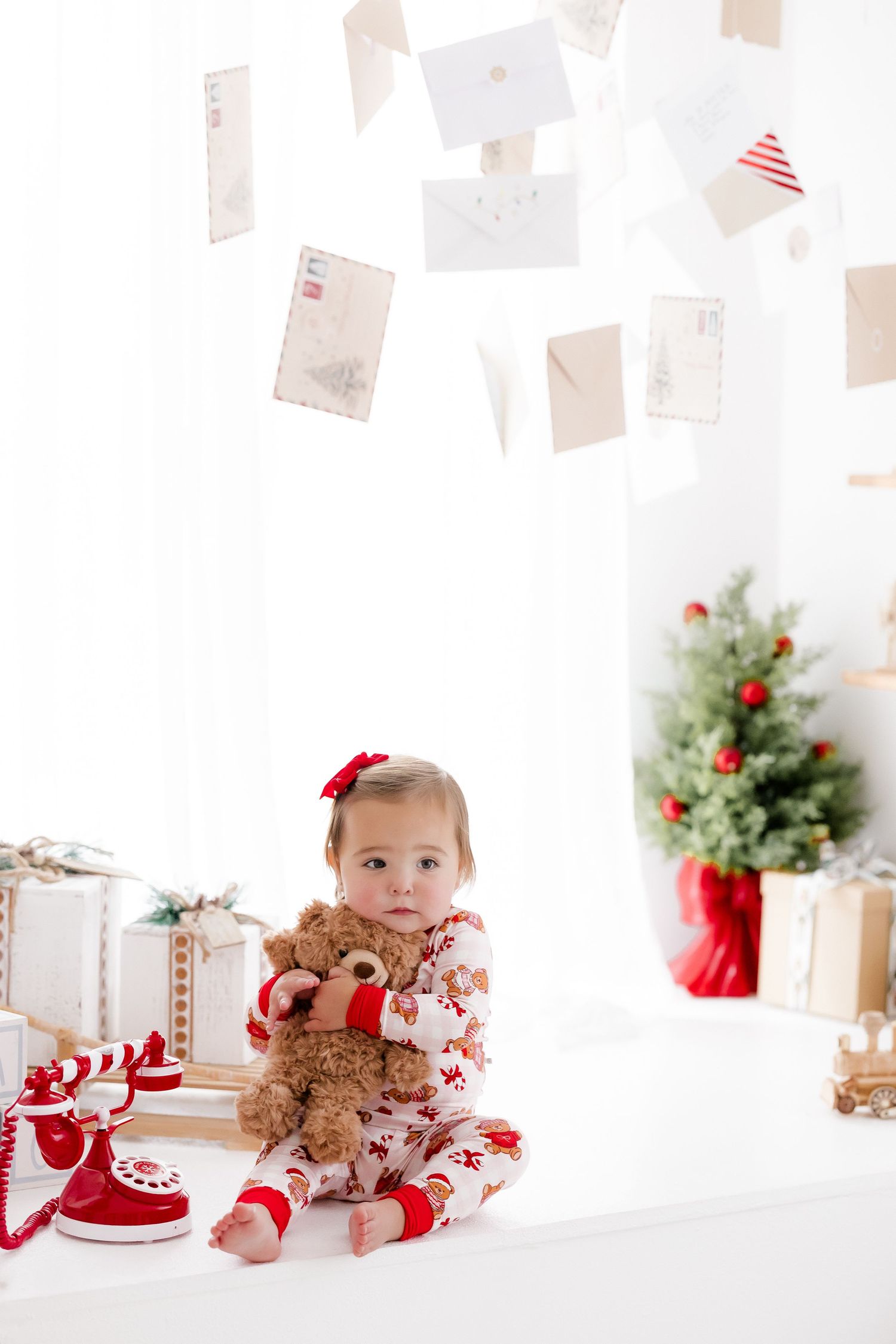 Cute baby girl in festive red and white pajamas holding a teddy bear near Christmas tree and gifts.