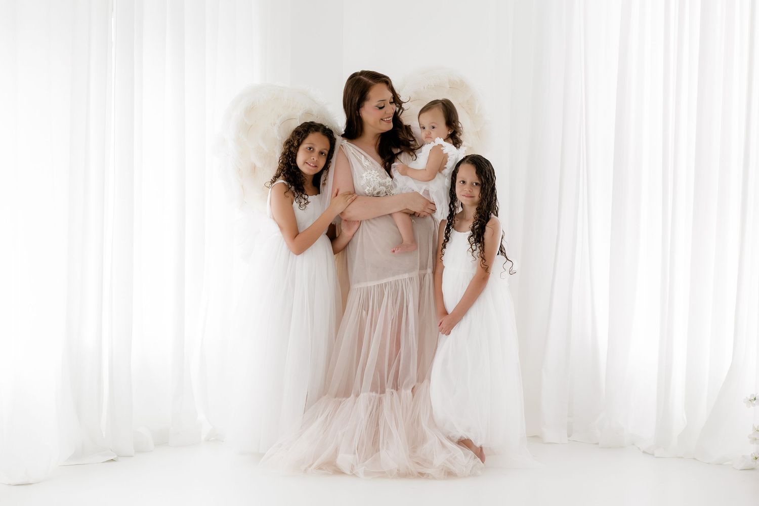 A mother in a pink gown holds her baby girl while two daughters in white dresses pose with angel wings.
