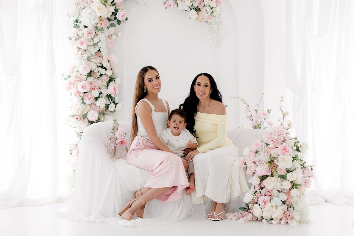 Three people in pastel dresses pose together in a white studio setting decorated with pink and white floral arrangements.