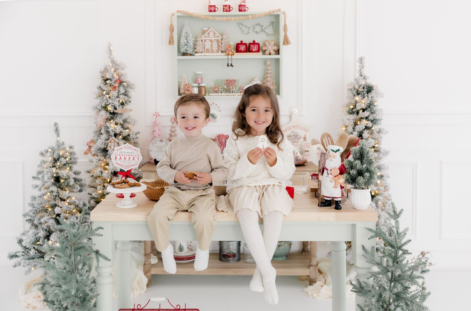 Two young children sit together on a white table surrounded by festive Christmas trees and holiday decorations in a bright studio.