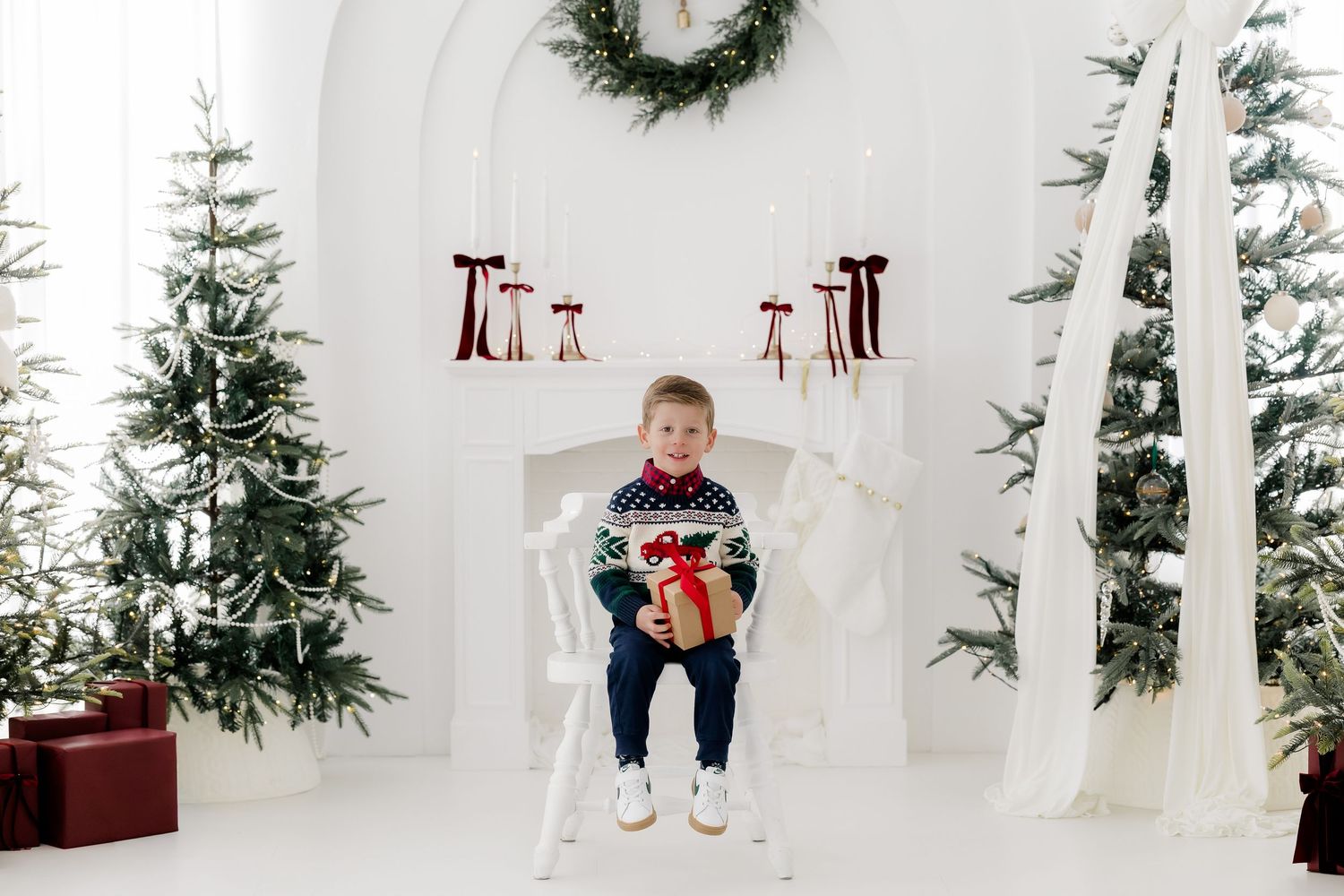 Young boy in holiday sweater sits on chair holding gift box surrounded by Christmas trees and white fireplace decor.