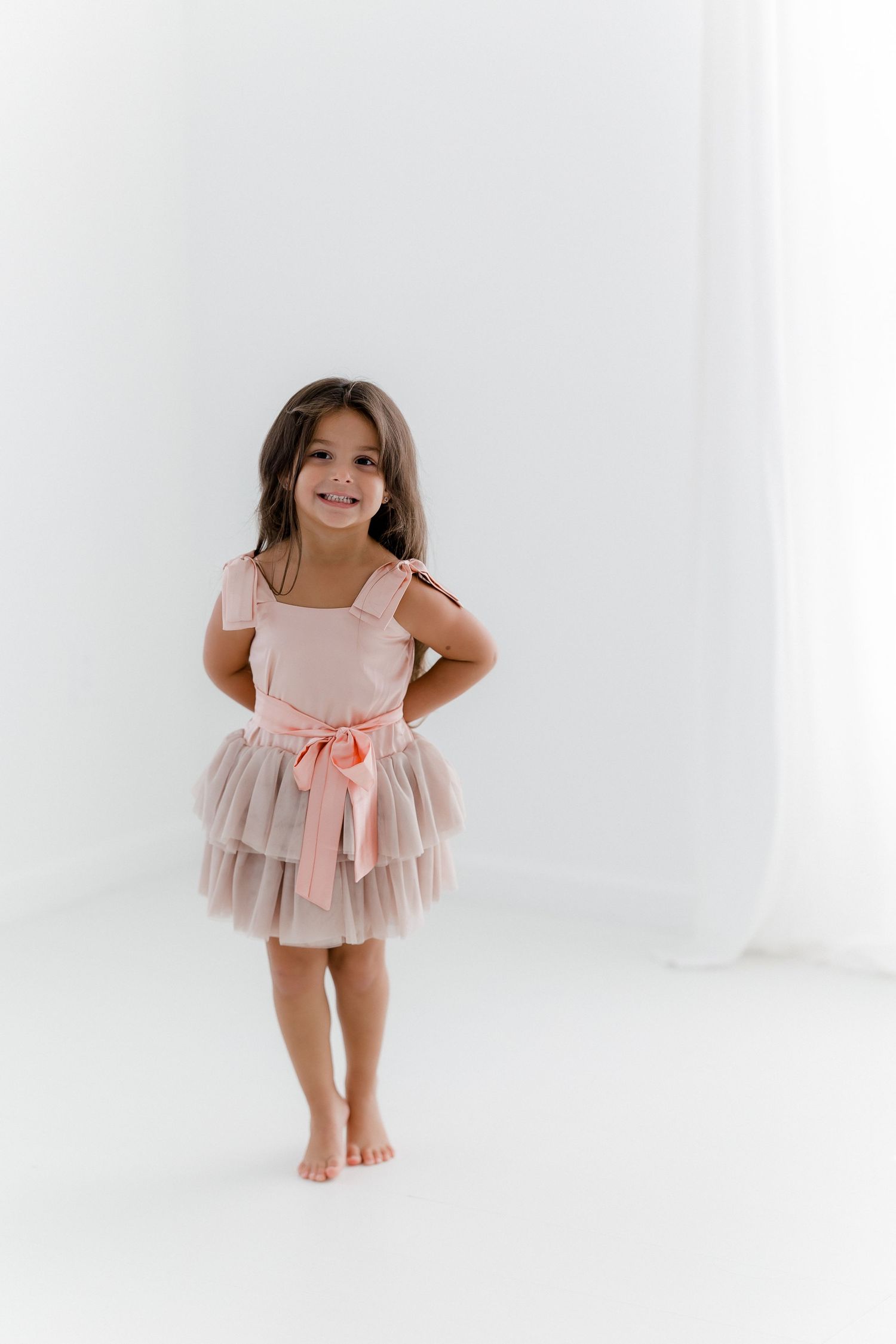 A child in a blush tulle dress with bow sash poses playfully in a bright studio setting.