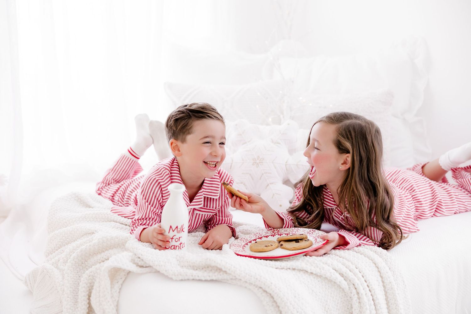 Siblings in matching pink pajamas share laughter and snacks while reading magazines on a cozy white bed. nj holiday mini sessions