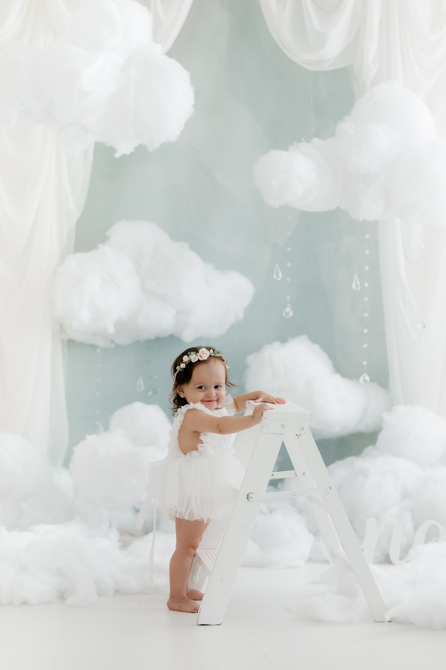 Adorable toddler girl in white tutu dress with floral crown standing by a ladder among fluffy white clouds.