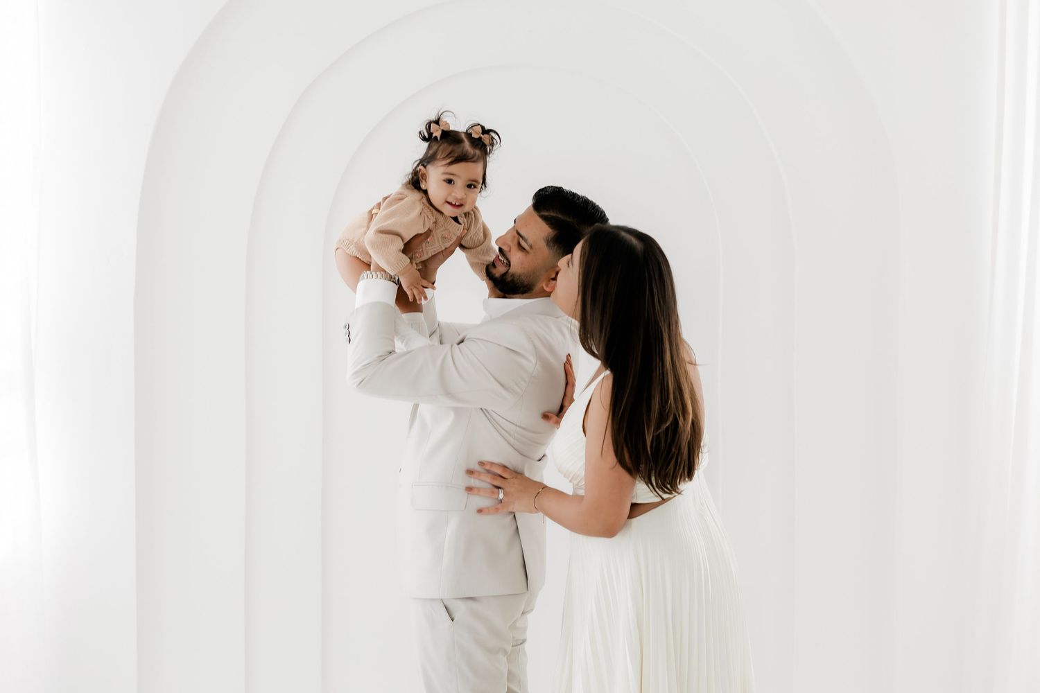 Mother and baby wearing white outfits embrace while posing against a bright white studio backdrop.