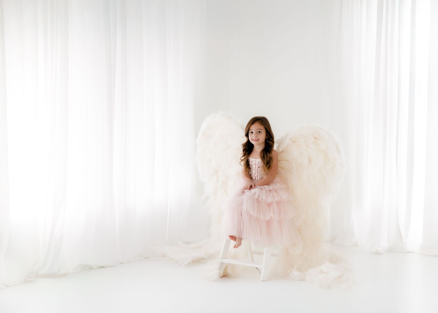 A bride in a white wedding dress poses in a bright, ethereal setting with soft lighting and white curtains.
