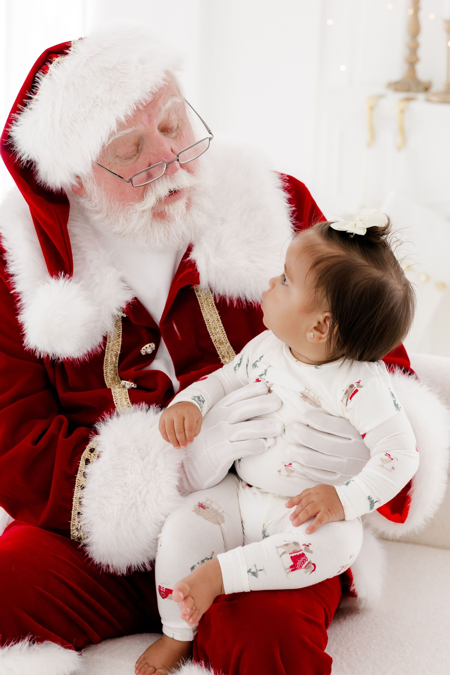 Santa Claus in red suit holds a baby in Christmas pajamas, sharing a tender holiday moment in a bright white setting.