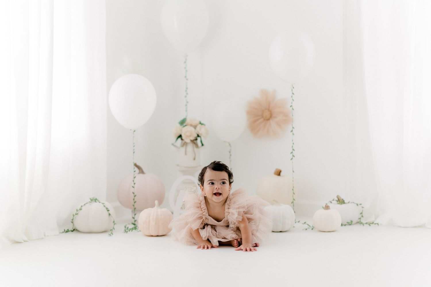 A baby sits on a white floor surrounded by blush pink balloons and floral decorations in a bright studio setting.