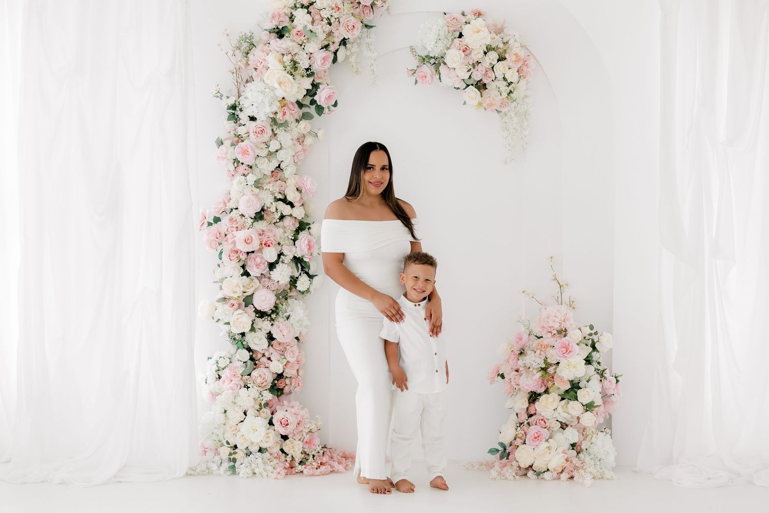 Elegant maternity photo featuring white gown against floral arch with pink and white roses on white backdrop.