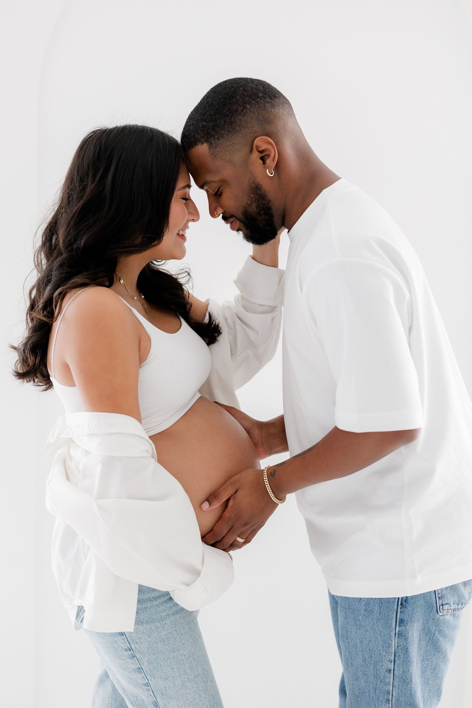 Intimate maternity photo series showing couple in white tops and jeans embracing against light background.