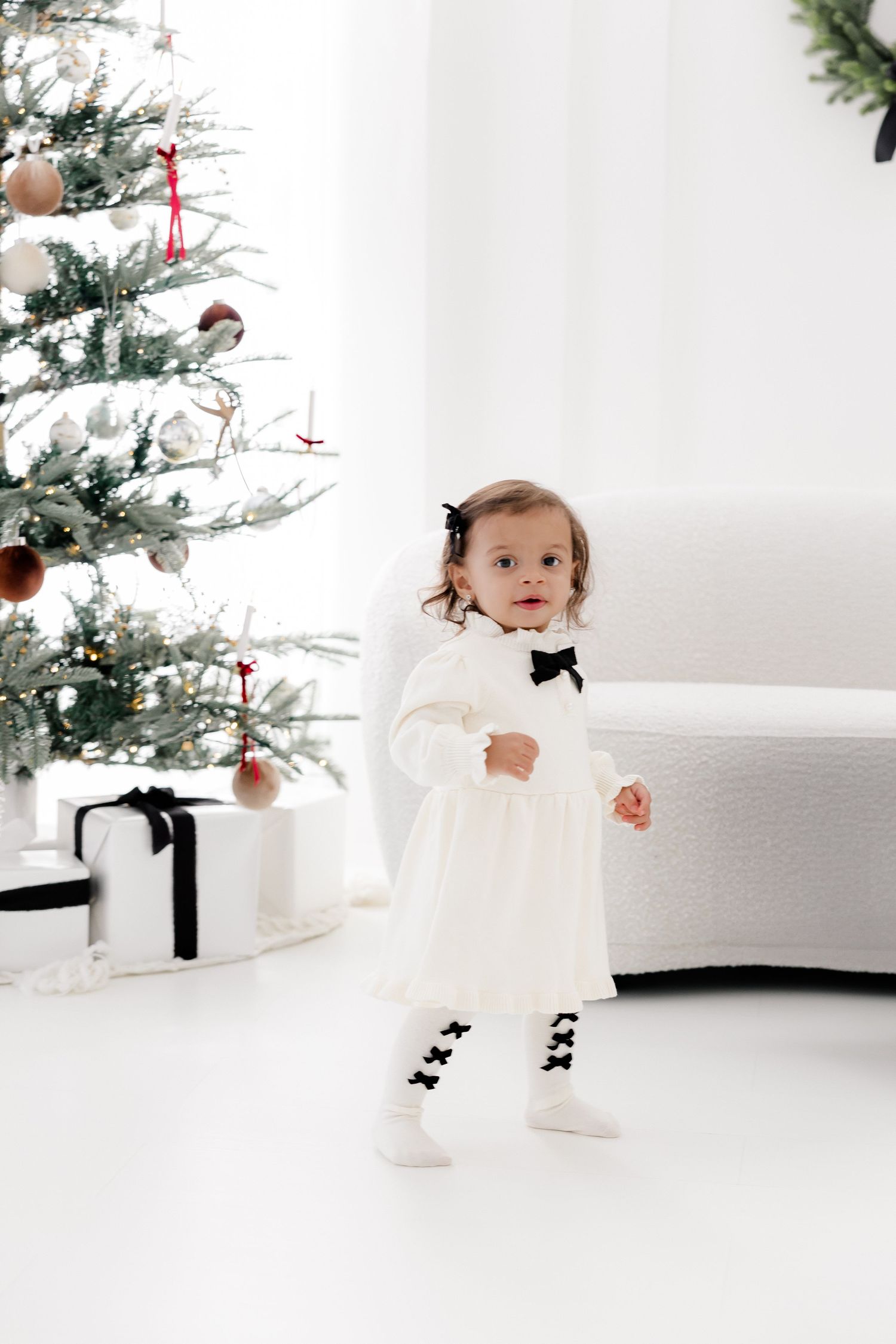 Small child in white dress poses near decorated Christmas tree in bright studio setting.