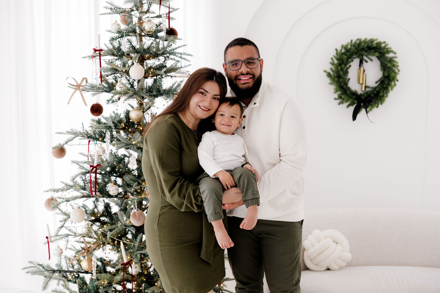 Family poses together near decorated Christmas tree and wreath in white room.
