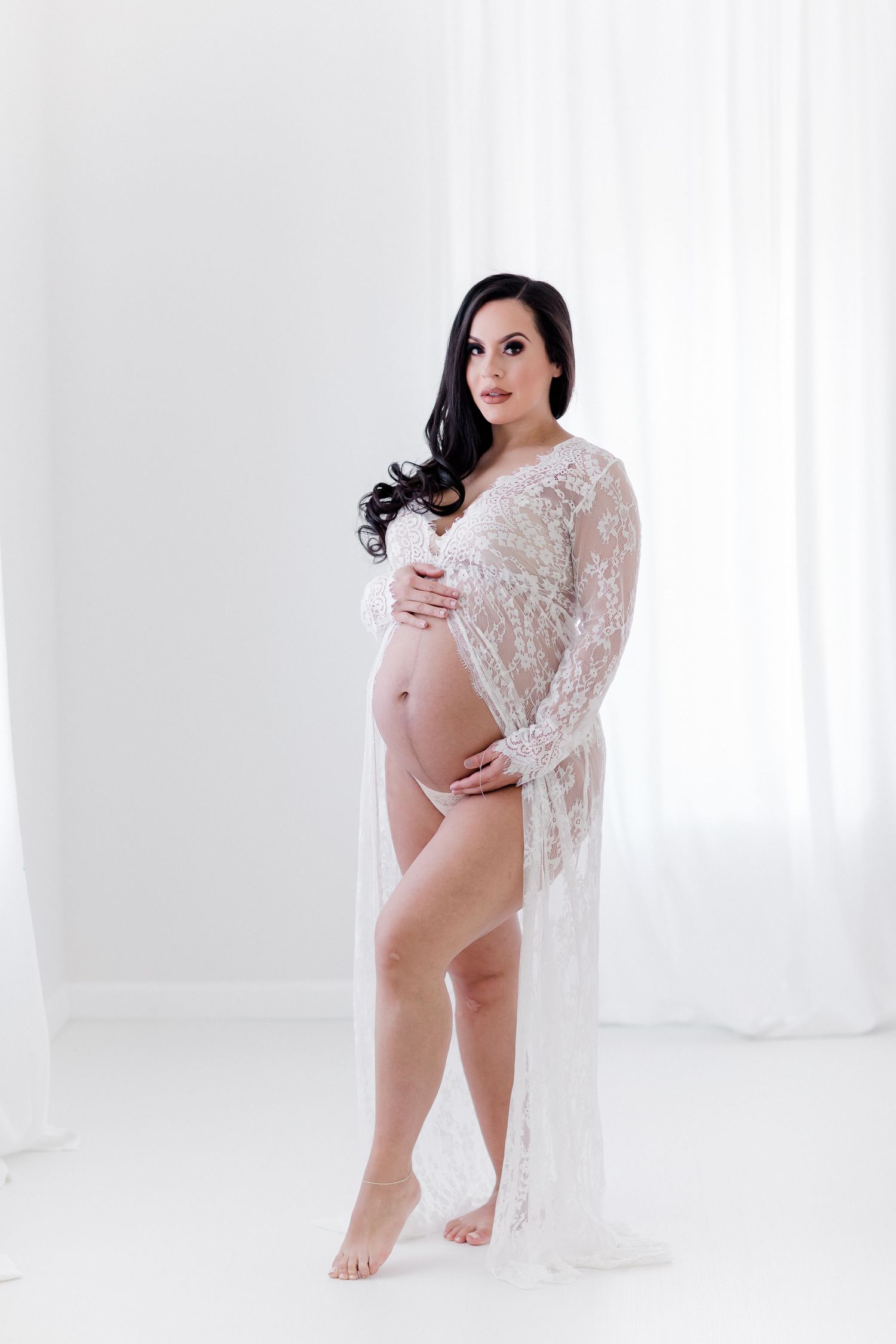 Graceful maternity photo with intricate white lace gown against pure white backdrop.