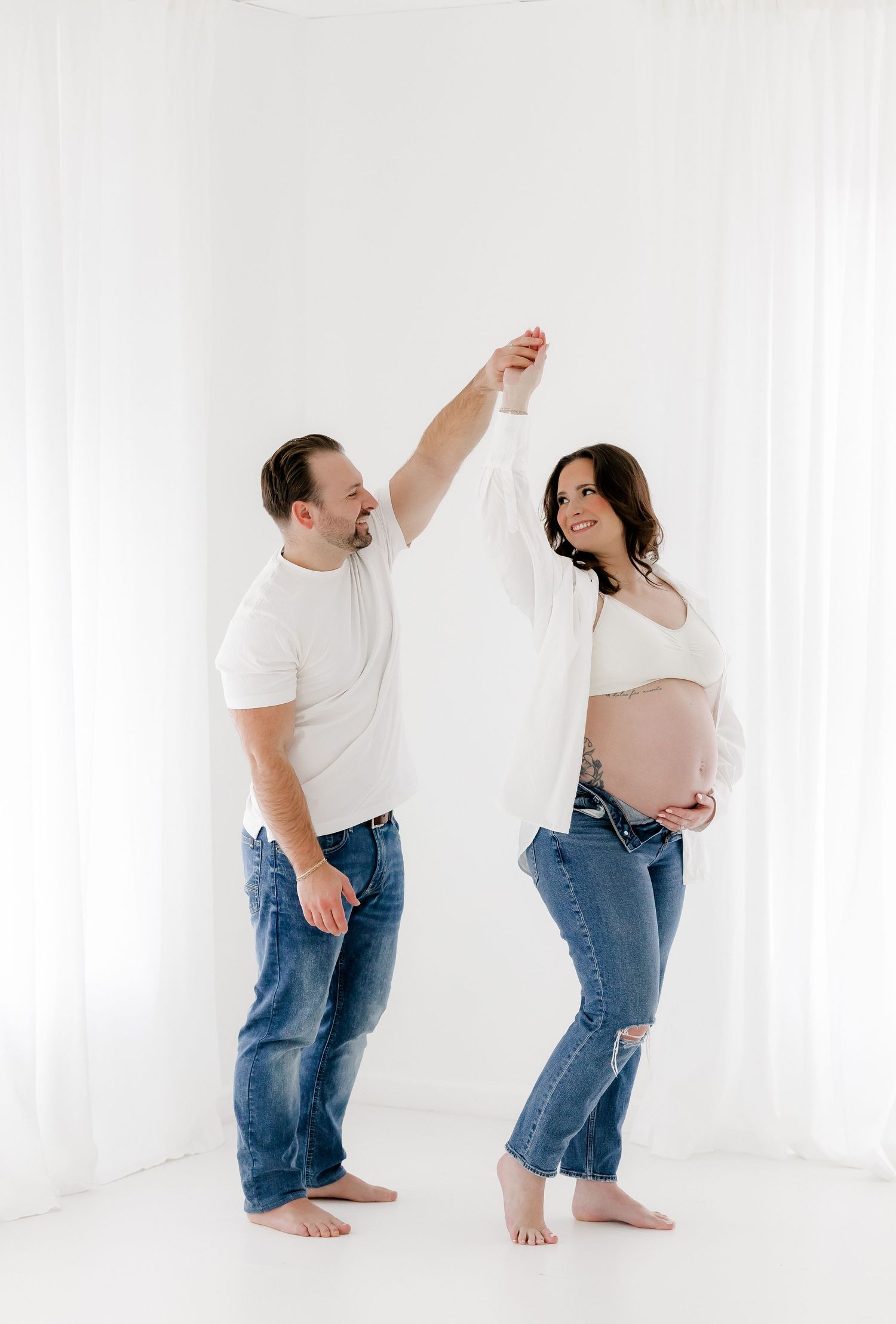 Expectant couple in matching white tops and jeans dance together against white backdrop in minimalist maternity photo.