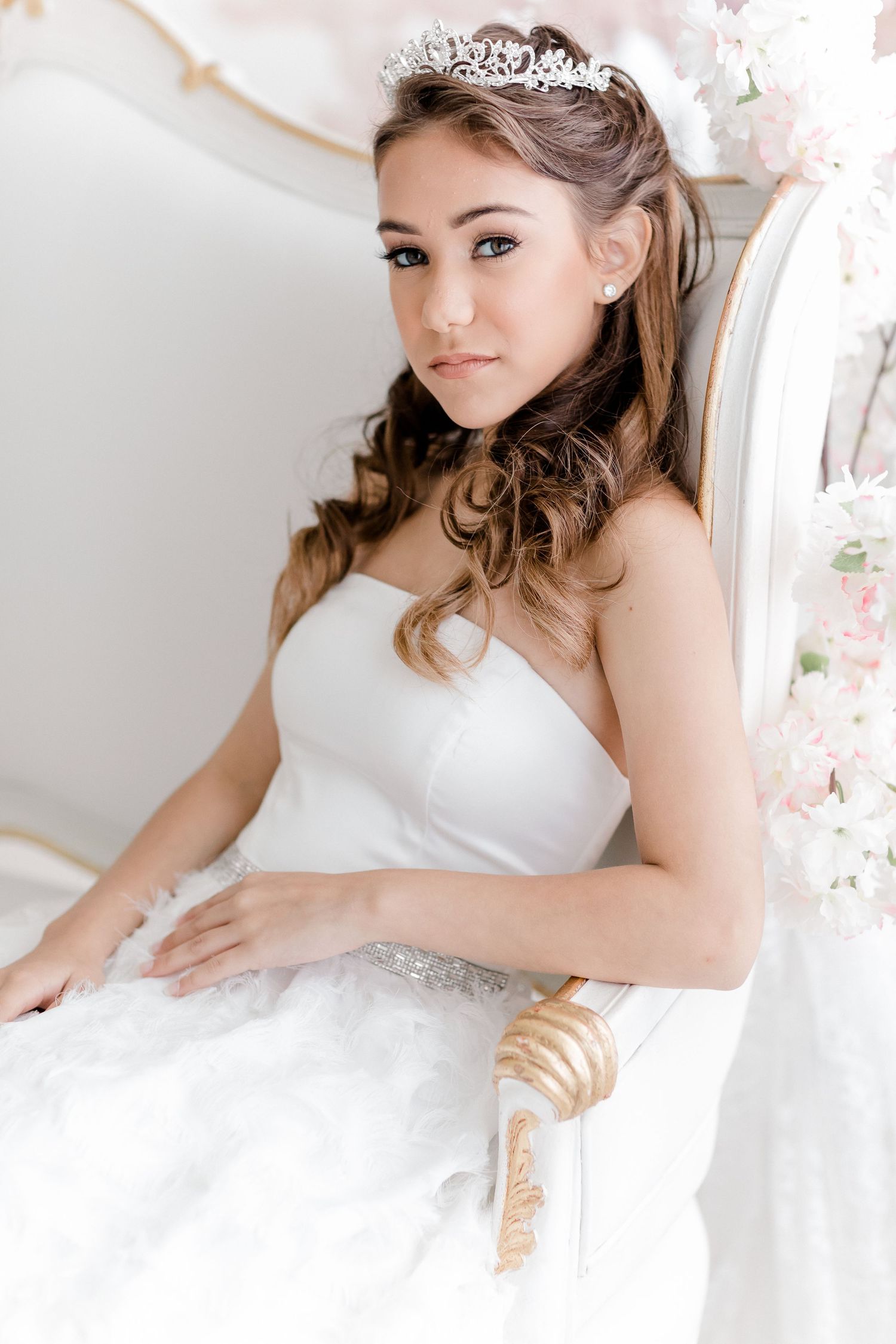 A bride in a white wedding gown with sparkly details poses elegantly on a white couch in a bright, airy setting.