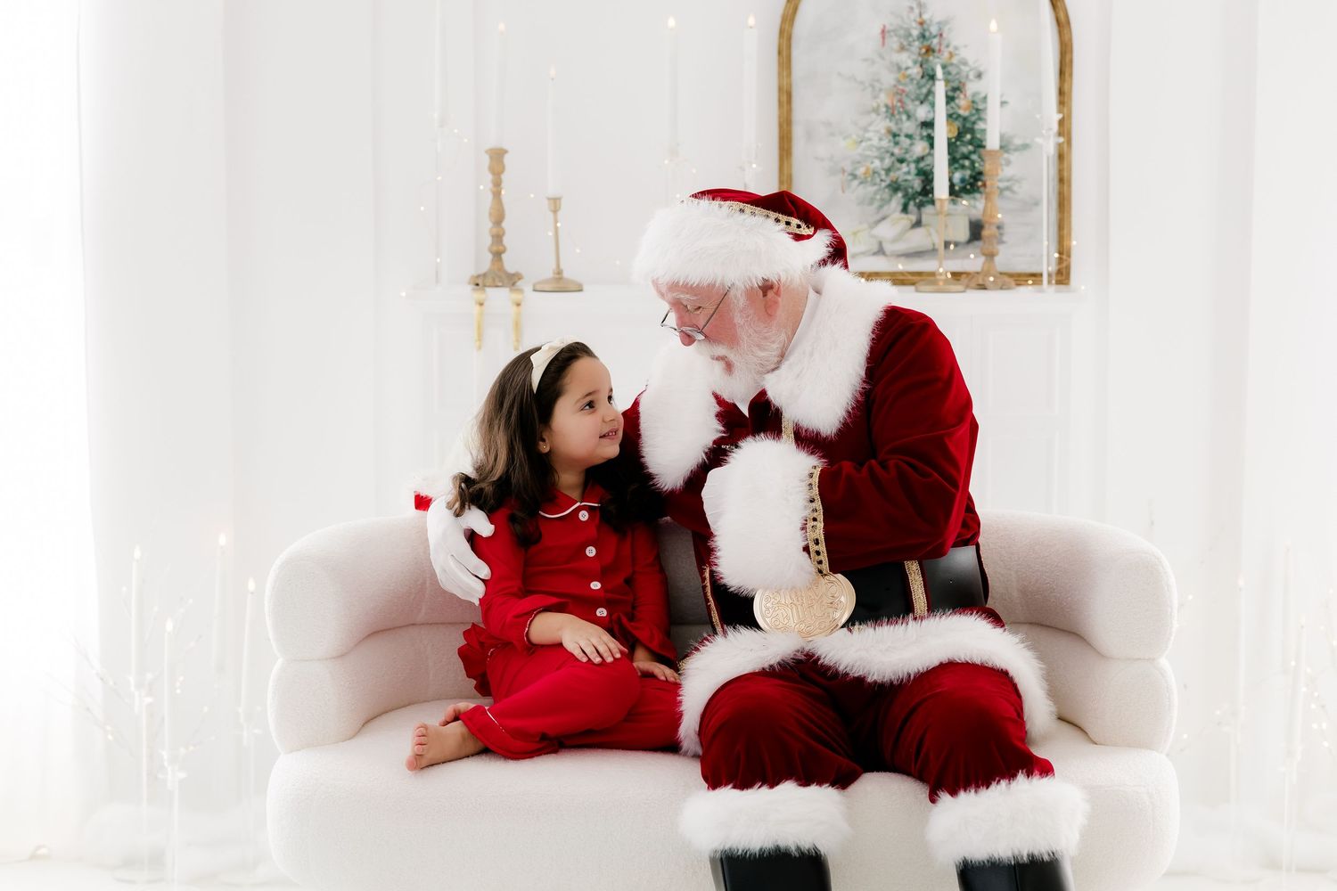 A young girl in a red dress sits with Santa Claus on a white sofa in a festive holiday photo setting.